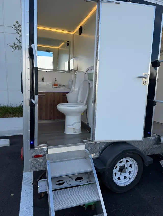 Open restroom trailer door with steps showing a toilet and wood vanity