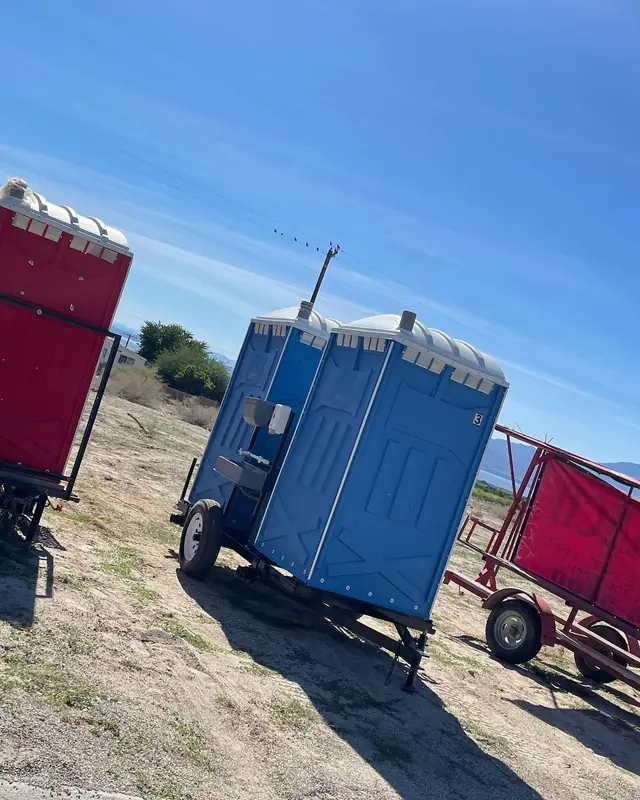 Two blue porta potties mounted on a small towable trailer at an open site